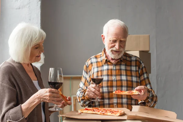 Coppia anziana che tiene pezzi di pizza e bicchieri di vino in una nuova casa — Foto stock