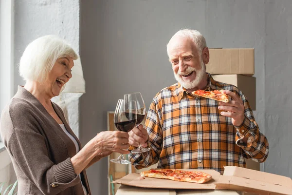 Laughing senior man holding piece of pizza and clinking glasses of wine with wife in new house — Stock Photo