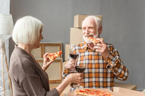 Rire couple âgé manger de la pizza et tenant des verres de vin dans une nouvelle maison — Photo de stock