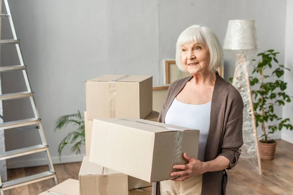 Cheerful senior woman holding cardboard box and looking away, moving concept — Stock Photo