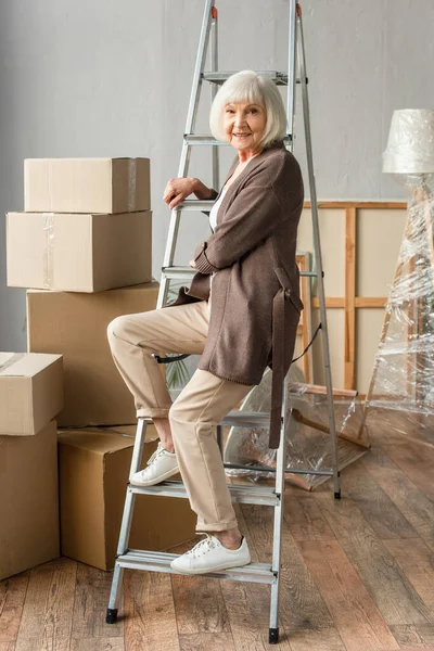 Вид в полный рост улыбающейся пожилой женщины, сидящей на лестнице с картонными коробками на заднем плане, движущаяся концепция — стоковое фото