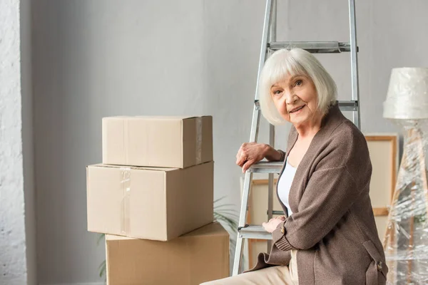 Donna anziana seduta sulla scala con scatole di cartone sullo sfondo, concetto in movimento — Foto stock