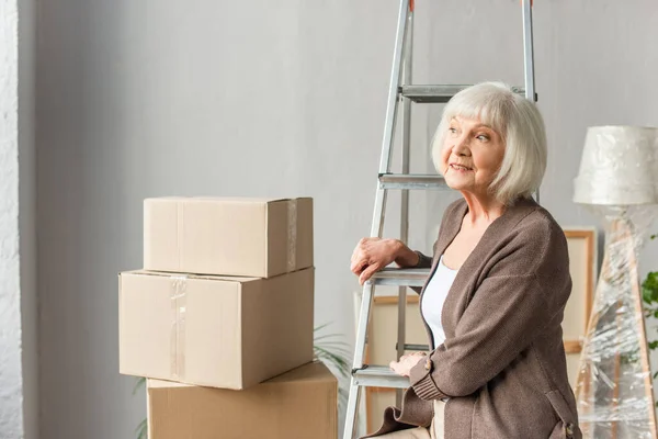 Улыбающаяся пожилая женщина сидит на лестнице и смотрит в сторону с картонными коробками на заднем плане, движущаяся концепция — стоковое фото