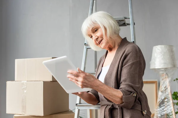 Senior woman sitting on ladder and using digital tablet with cardboard boxes on background, moving concept — Stock Photo