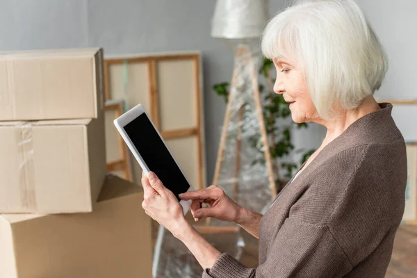 Side view of senior woman holding digital tablet with blank screen with cardboard boxes on background, moving concept — Stock Photo
