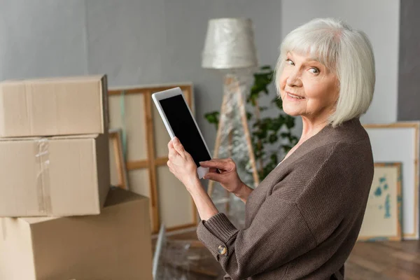 Senior femme tenant tablette numérique avec écran blanc et regardant en arrière avec des boîtes en carton sur fond, concept mobile — Photo de stock