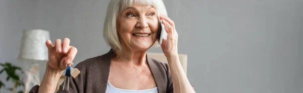 Panoramic shot of happy senior woman talking on phone and holding keys in new house, moving concept — Stock Photo