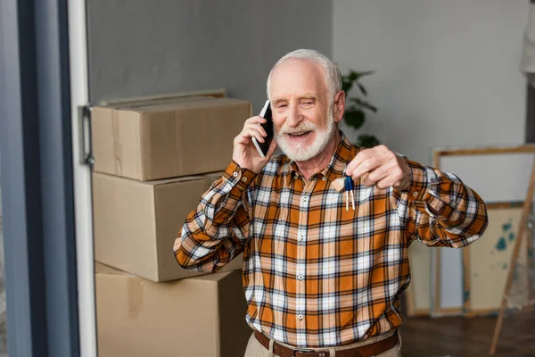 Hombre mayor feliz hablando por teléfono y sosteniendo las llaves en la nueva casa, concepto en movimiento - foto de stock
