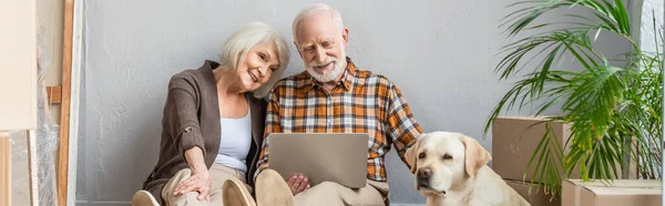 Plan panoramique de heureux couple de personnes âgées en utilisant ordinateur portable assis sur le sol et mari chien caressant couché près — Photo de stock