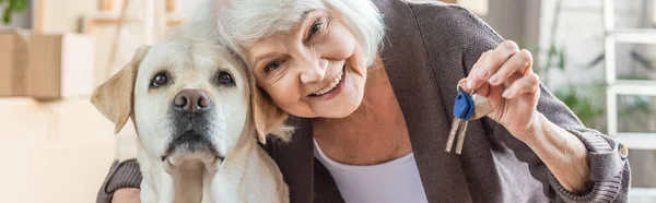 Tiro panorâmico de sorrir mulher sênior abraçando cão e segurando chaves, conceito em movimento — Fotografia de Stock