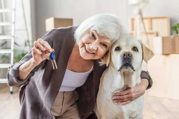 Smiling senior woman embracing dog and holding keys, moving concept — Stock Photo
