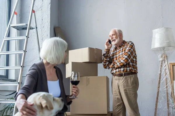 Mulher sênior abraçando cão e segurando copo de vinho, enquanto o homem falando no telefone — Fotografia de Stock