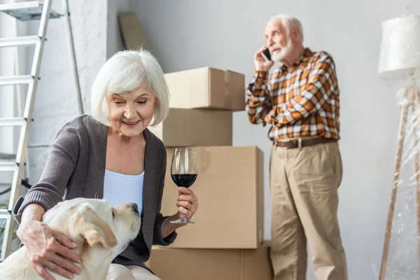 Senior mujer abrazando perro y sosteniendo vaso de vino mientras hombre hablando por teléfono - foto de stock