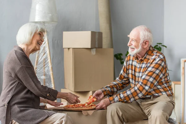 Rire couple âgé prendre des morceaux de pizza dans la maison neuve et boîtes en carton sur fond — Photo de stock