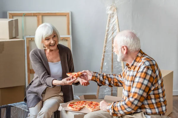 Riant senior homme donnant femme morceau de pizza dans la nouvelle maison et boîtes de carton sur fond — Photo de stock