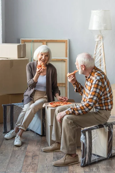 Ganzkörperansicht des lachenden Seniorehepaares beim Pizza essen im neuen Haus und Pappkartons im Hintergrund — Stockfoto