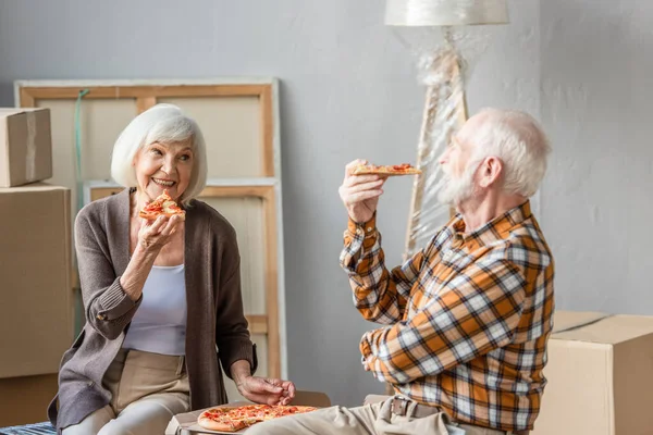 Rire couple de personnes âgées manger de la pizza dans une nouvelle maison et boîtes en carton sur fond — Photo de stock