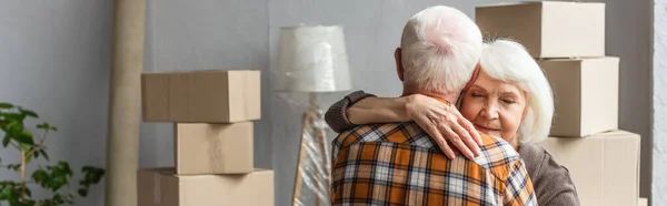 Panoramic shot of senior woman with closed eyes hugging husband in new house, moving concept — Stock Photo