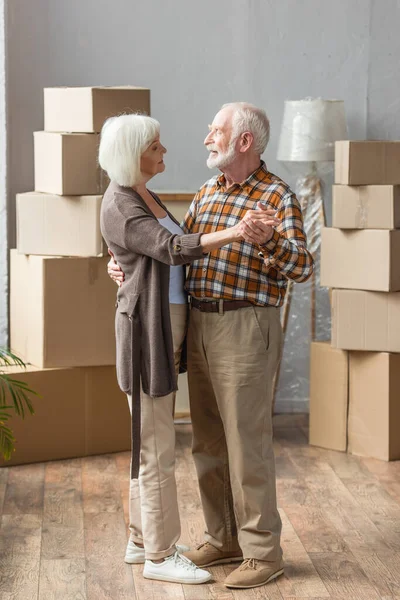 Full length view of cheerful senior couple dancing in new house with cardboard boxes on background — Stock Photo