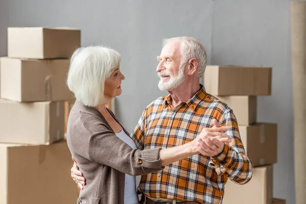 Cheerful senior couple dancing in new house with cardboard boxes on background — Stock Photo