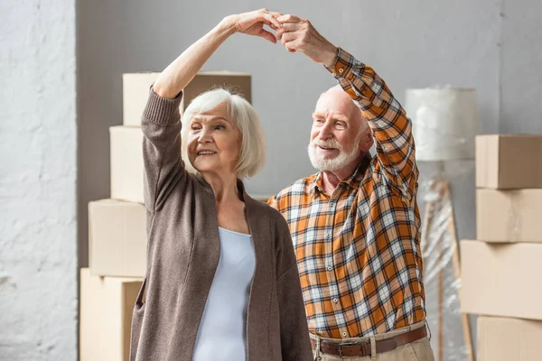 Cheerful senior couple dancing in new house with cardboard boxes on background — Stock Photo