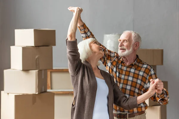 Cheerful senior couple dancing in new house with cardboard boxes on background — Stock Photo