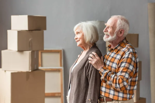 Cheerful husband hugging wife shoulders in new house with cardboard boxes on background — Stock Photo