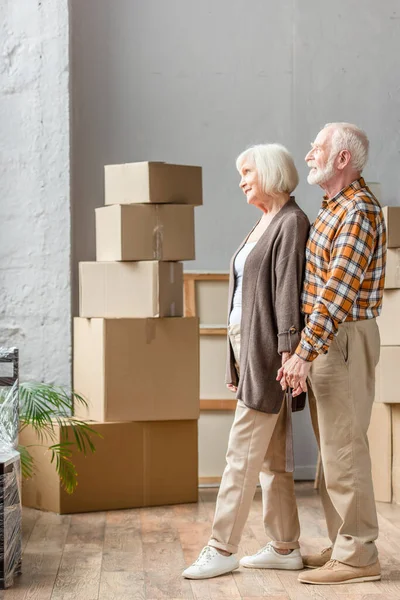 Pareja de ancianos tomados de la mano y mirando en la ventana en la nueva casa, concepto móvil - foto de stock