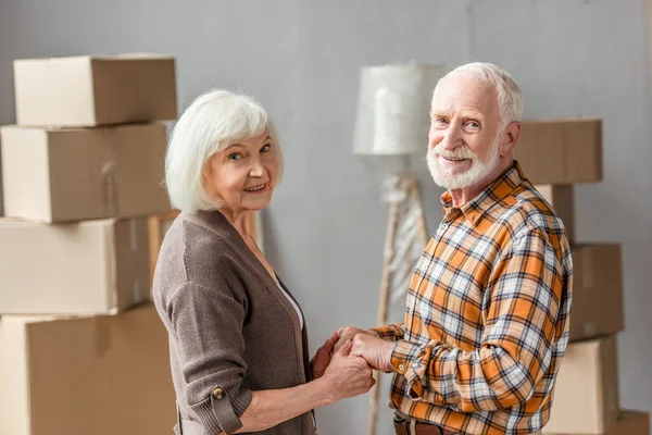 Senior couple holding hands and looking at camera in new house, moving concept — Stock Photo