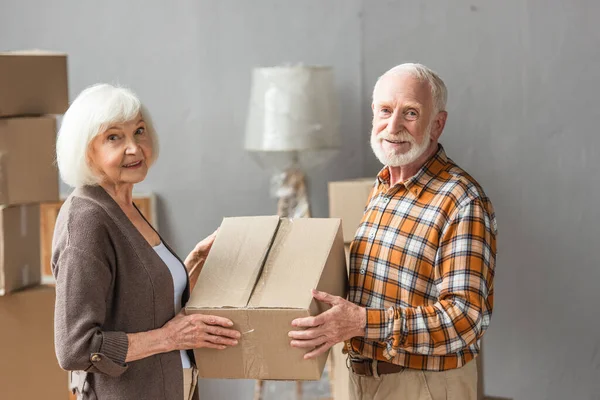 Senior couple holding cardboard box and looking at camera in new house, moving concept — Stock Photo