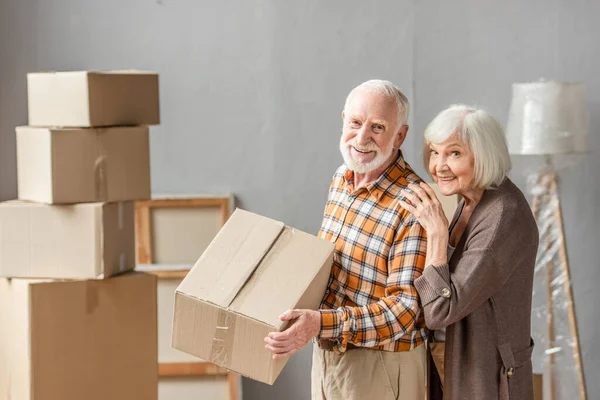 Senior man holding cardboard box and wife leaning on shoulder in new house, moving concept — Stock Photo