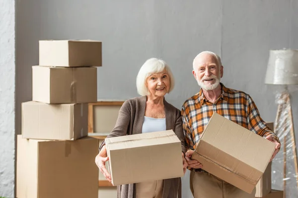 Пожилая пара держит картонные коробки и смотрит на камеру в новом доме, движущаяся концепция — стоковое фото