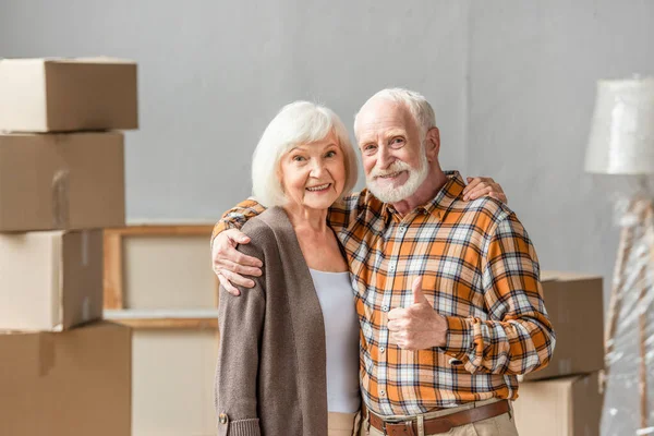 Senior couple embracing and man showing thump up in new house, moving concept — Stock Photo