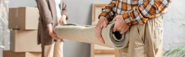 Panoramic shot of senior couple rolling carpet in new house, moving concept — Stock Photo