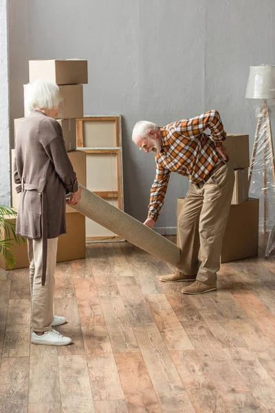 Full length of senior man suffering from backache while rolling carpet with wife — Stock Photo