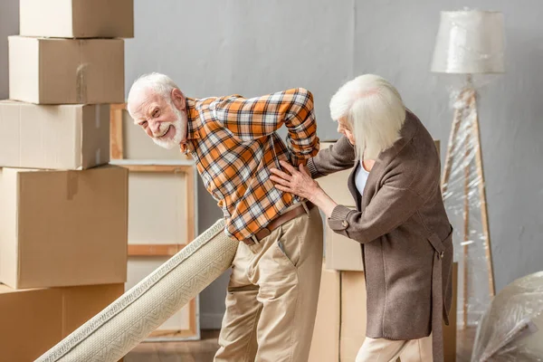 Senior man suffering from backache and holding carpet while wife trying help — Stock Photo