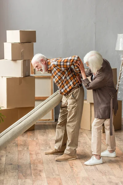 Senior man suffering from backache and holding carpet while wife trying help — Stock Photo