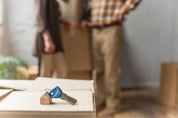 Keys lying on cardboard box on foreground and blurred view of senior couple, moving concept — Stock Photo