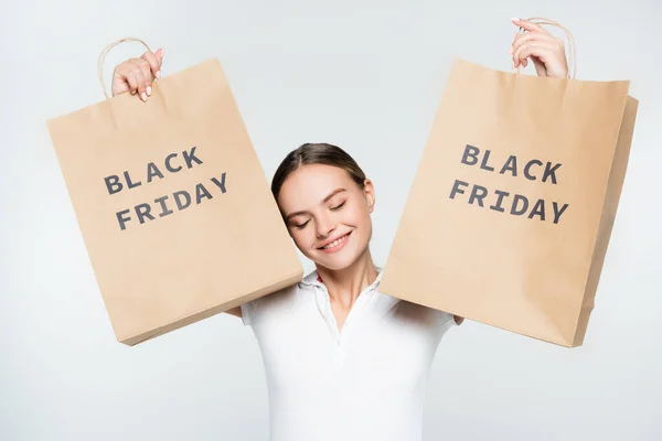 Joven mujer alegre con los ojos cerrados sosteniendo bolsas de compras con letras de viernes negro aislado en blanco - foto de stock