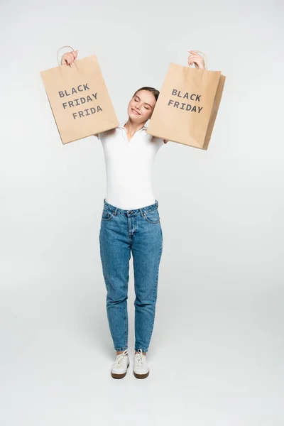 Mujer joven con los ojos cerrados sosteniendo bolsas de compras con letras de viernes negro en blanco - foto de stock