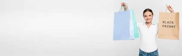 Panoramic shot of pleased young woman holding shopping bags with black friday lettering on white — Stock Photo