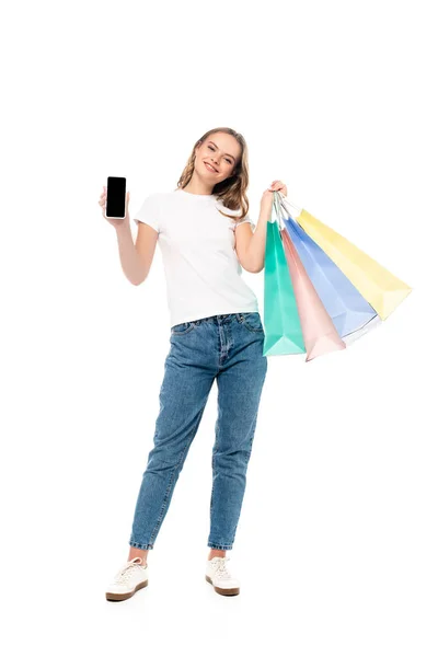 Mujer joven satisfecha sosteniendo teléfono inteligente con pantalla en blanco cerca de coloridas bolsas de compras aisladas en blanco - foto de stock