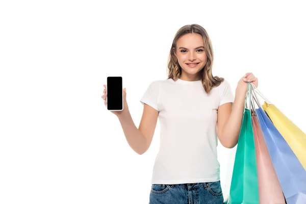 Freudige junge Frau hält Smartphone mit leerem Bildschirm in der Nähe bunter Einkaufstüten isoliert auf weiß — Stockfoto