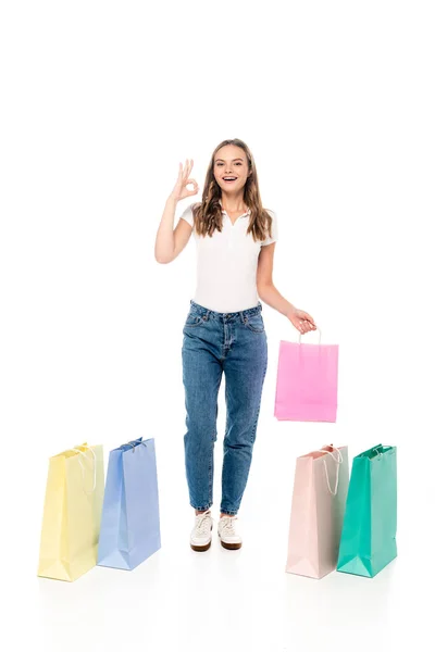 Complacida y joven mujer mostrando signo ok cerca de coloridas bolsas de compras aisladas en blanco - foto de stock