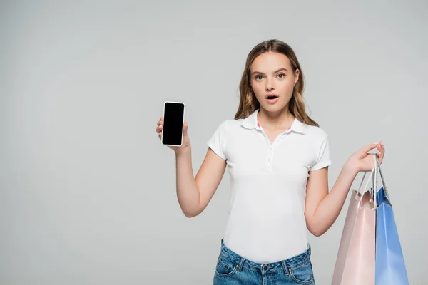 Donna scioccata con smartphone con schermo bianco e borse della spesa isolate sul grigio — Foto stock