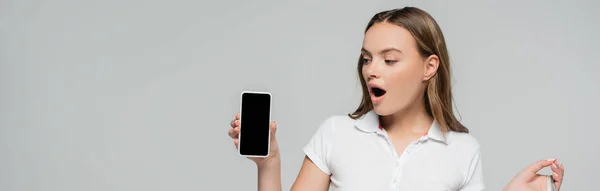 Concepto panorámico de mujer sorprendida mirando el teléfono inteligente con pantalla en blanco aislado en gris - foto de stock