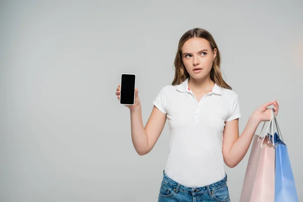 Pensive woman holding smartphone with blank screen and shopping bags isolated on grey — Stock Photo