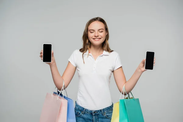 Joyful woman with closed eyes holding mobile phones with blank screen and shopping bags isolated on grey, black friday concept — Stock Photo