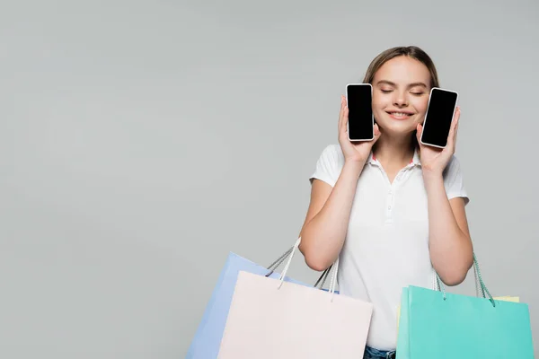 Задоволена жінка з закритими очима тримає мобільні телефони з порожнім екраном і сумки для покупок ізольовані на сірій, чорній п'ятниці концепції — стокове фото