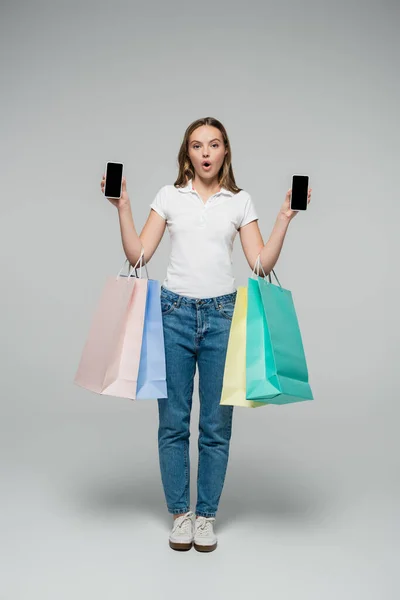 Шокированная женщина держит мобильные телефоны с чистым экраном и сумки для покупок на серой, черной пятницы концепции — стоковое фото